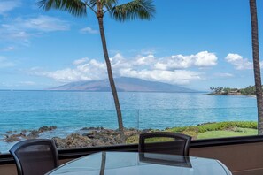 A view from a window overlooking a body of water with a mountain in the distance, a chair, and a table. - Makena Surf, #f-206 2 Bedroom Condo (Kihei)