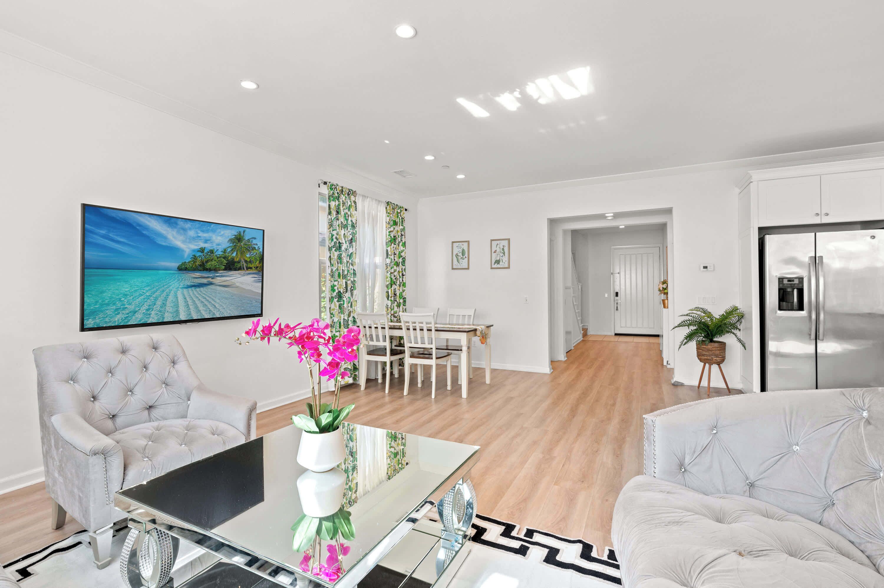 Una sala de estar moderna con un televisor de pantalla plana, dos sillones tapizados, una mesa de centro de vidrio y un comedor con una mesa y sillas blancas.