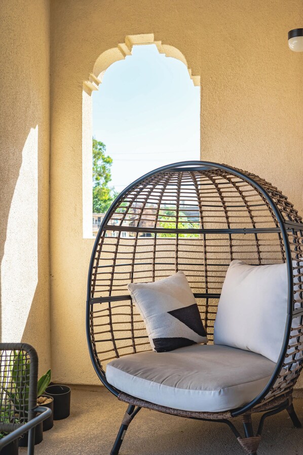 A round, wicker chair with cushions, positioned on a balcony with a view of a building and trees.