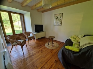 Ein Zimmer mit Holzboden, einem Fernseher auf einem weißen Schrank, einem Korbstuhl, einem runden Tisch und einem Sofa mit Kissen.