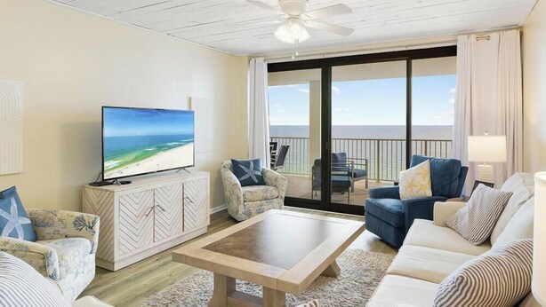 A living room with a TV, a ceiling fan, a balcony with a view of the ocean, and a large window. - Windward Pointe 1102 by Liquid Life (Orange Beach)
