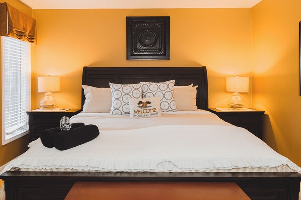 Una cama bien hecha con una almohada de bienvenida, dos lámparas de noche y una obra enmarcada sobre la cama.