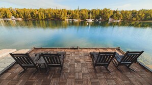 Una terraza de madera con sillas que da a un lago tranquilo, rodeado de árboles y casas.