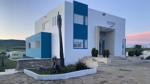 A modern building with a white and blue facade, stone-paved courtyard, and a view of the surrounding landscape. - Guitar Palace (Bni Quolla)