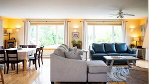 Uma sala de estar com um sofá cinza, um sofá azul, uma mesa de jantar com cadeiras e janelas grandes com cortinas.