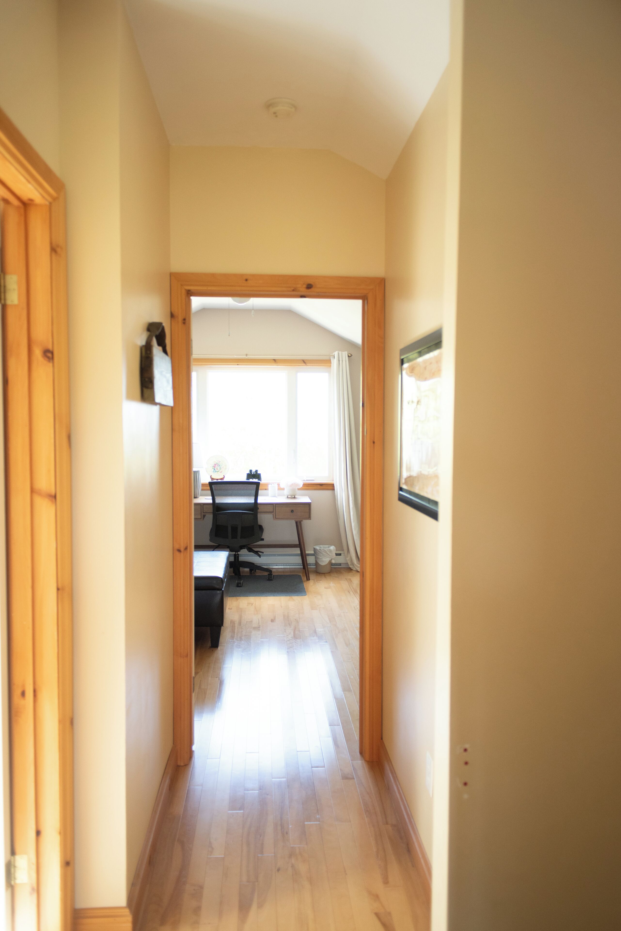 Un couloir étroit, doté d’un plancher et de murs en bois, mène à une pièce où se trouvent un bureau, une chaise et une fenêtre avec des rideaux.