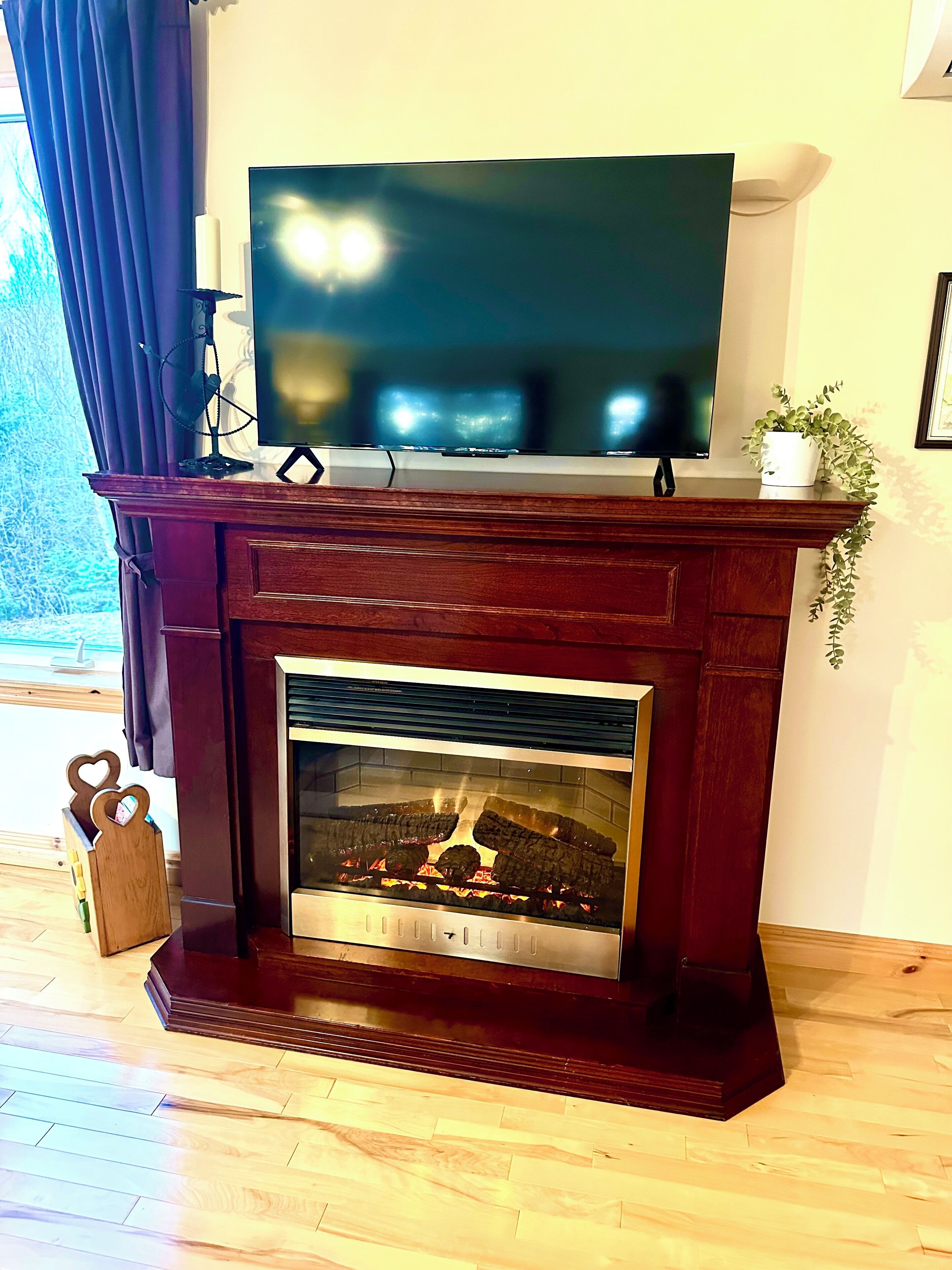 Una chimenea con un televisor encima, una planta sobre la repisa y un banco de madera con un portalápices con forma de corazón.