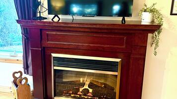 Una chimenea con un televisor encima, una planta sobre la repisa y un banco de madera con un portalápices con forma de corazón.