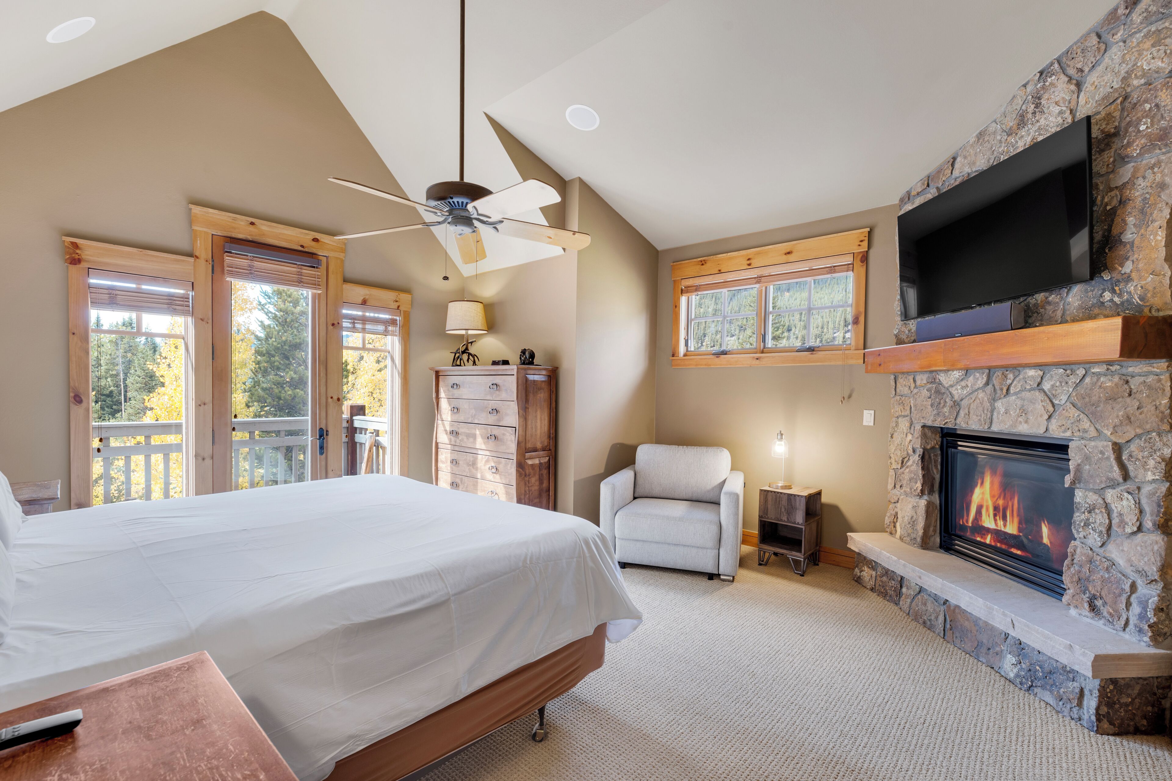 A bedroom with a large bed, a stone fireplace, a TV mounted on the wall, and a ceiling fan.