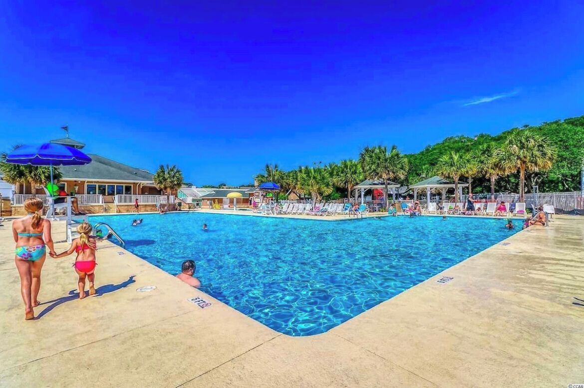 A swimming pool with lounge chairs and umbrellas, surrounded by palm trees and a building with a sloped roof.