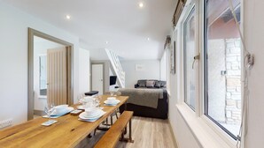 A dining area with a wooden table set for four, a sofa, and a large window with a view of a stone wall.