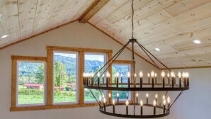 A room with a wooden ceiling, a chandelier with multiple lights, and three large windows offering a view of a mountainous landscape.