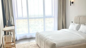 A bedroom with a large bed, a standing heater, a small table, and a pair of slippers on a rug.
