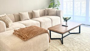 A modern living room with a beige sectional sofa, a wooden coffee table, a fluffy rug, and a large window with curtains.