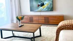 A modern living room with a wooden coffee table, a sofa, a TV mounted on the wall, and a large window with curtains.
