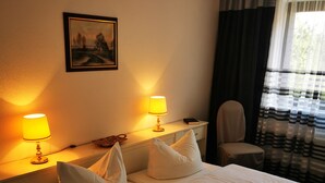 Une chambre d’hôtel avec un lit, deux lampes de chevet, une chaise, un tableau au mur et une fenêtre avec des rideaux.