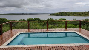 A wooden deck with a swimming pool, surrounded by lush greenery and a view of a lake.