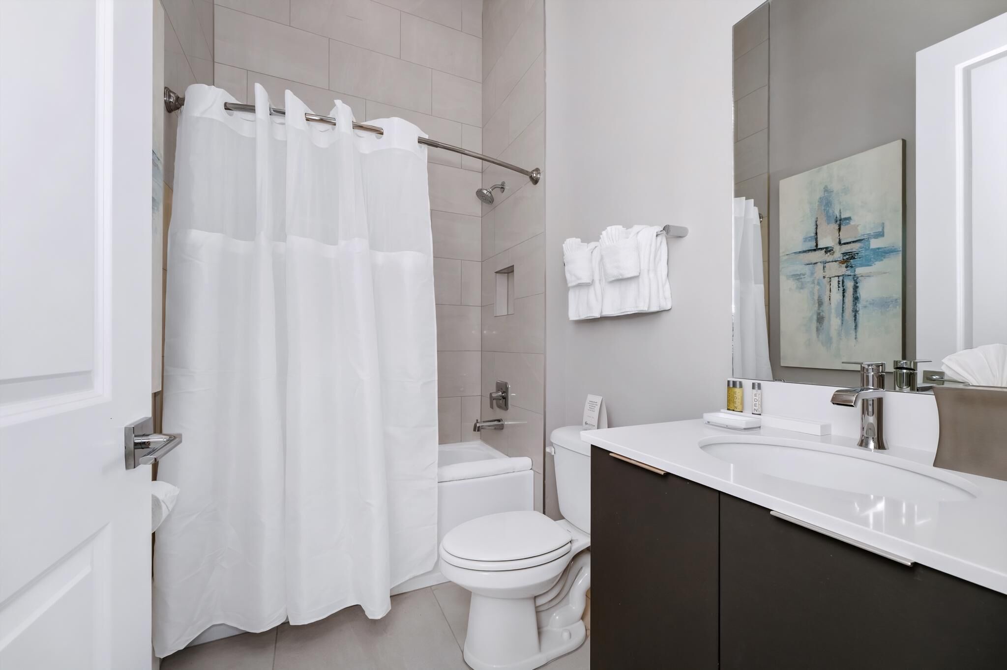 A modern bathroom with a shower curtain, toilet, sink, and a painting on the wall.
