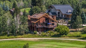A large, multi-story house with a prominent balcony, surrounded by lush greenery and a well-maintained lawn.
