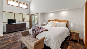 A spacious bedroom with a large bed, a wooden dresser, a television, and a window with a view.