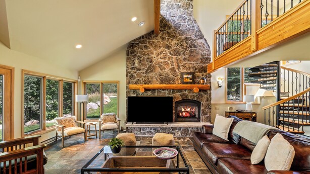 A spacious living room with a stone fireplace, a large flat-screen TV, leather sofas, and a wooden staircase.