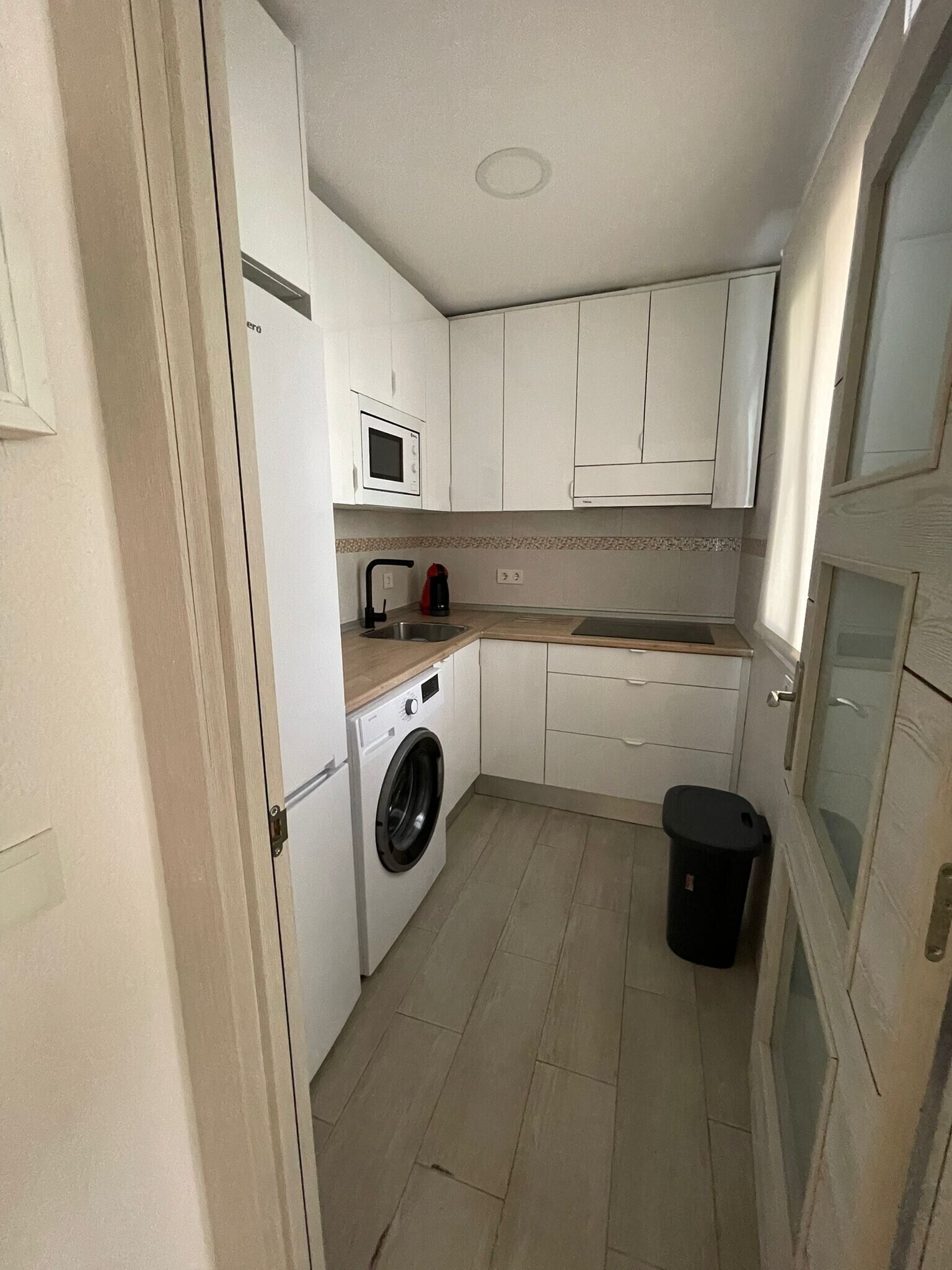 Una cocina compacta con armarios blancos, lavadora, microondas y un cubo de basura.