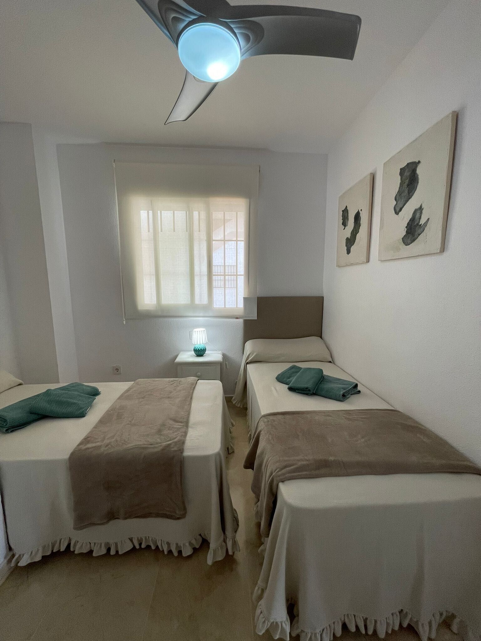 Een slaapkamer met twee bedden, een plafondventilator, een raam met zonwering en kunst aan de muur.