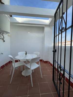 A patio with a round table and white chairs, a potted plant, and a view through a metal gate.