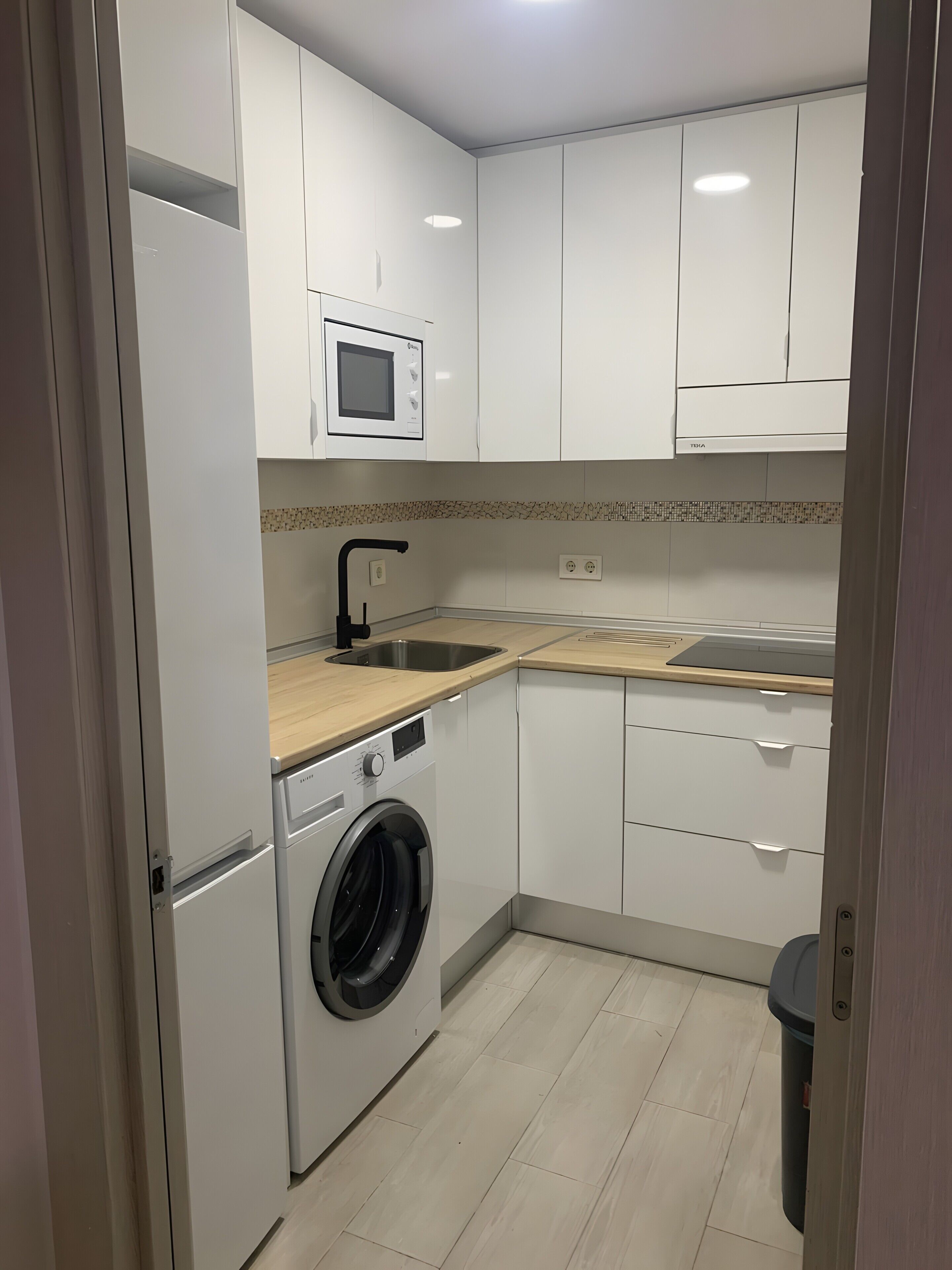 Cocina compacta con armarios blancos, microondas integrado, lavadora y cubo de basura.