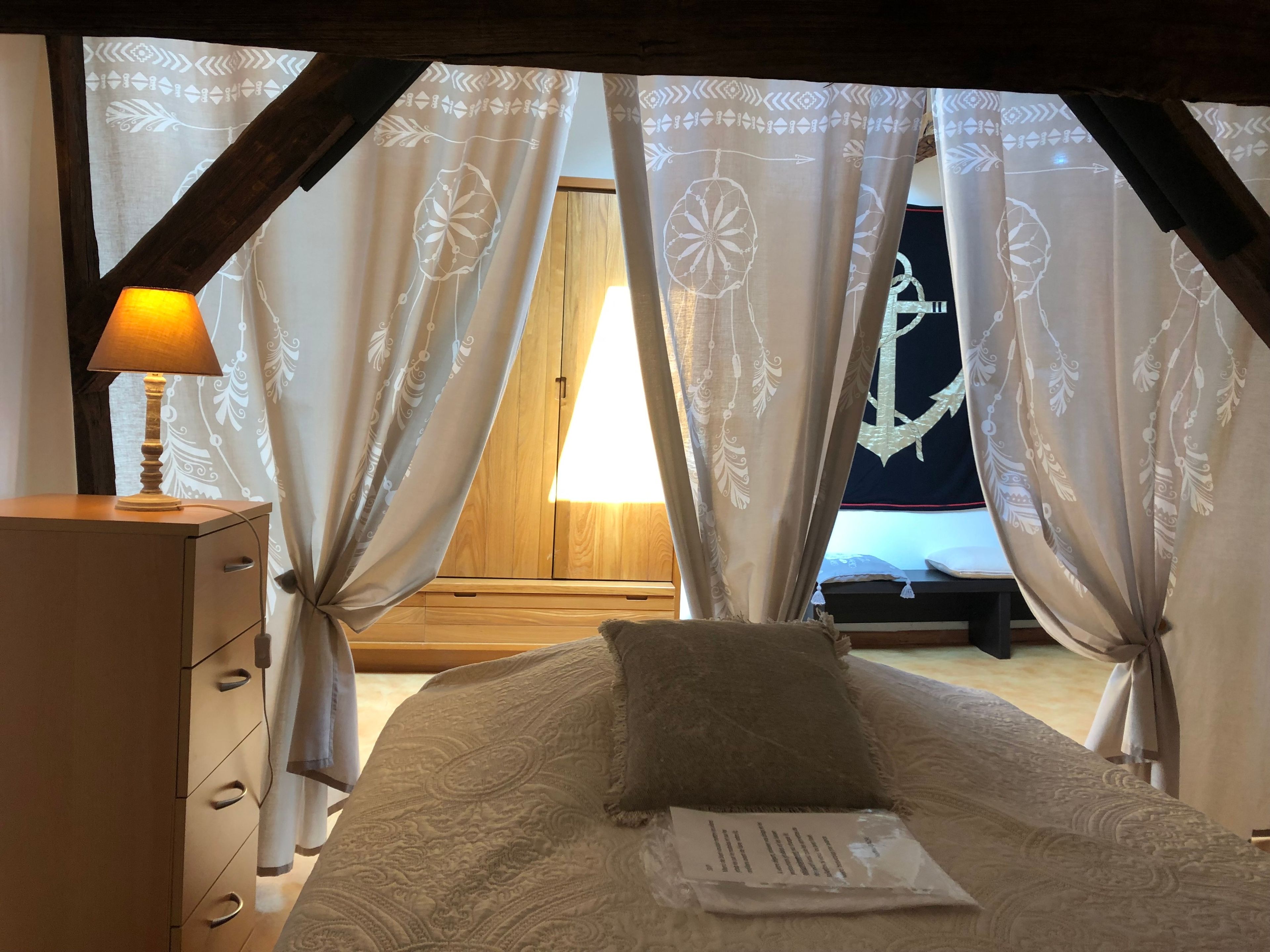 Quarto com cama, mesa de cabeceira, abajur e cortinas finas com padrão.