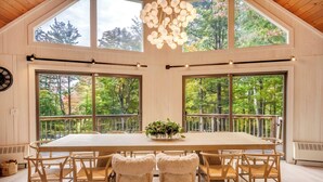 Un vaste espace dédié à la salle à manger, avec une grande table et des chaises, de grandes fenêtres offrant une vue sur les arbres et un lustre moderne.