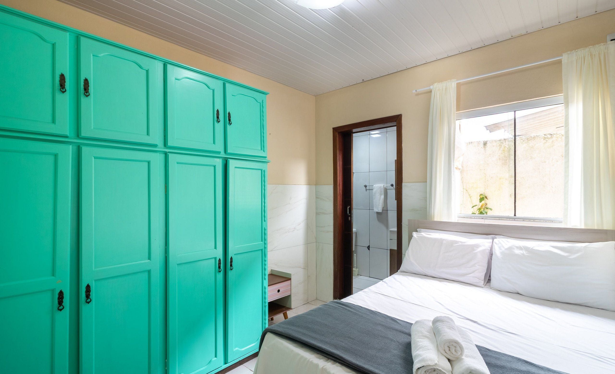 A hotel room with a bed, a large teal wardrobe, a window with curtains, and a small bedside table.