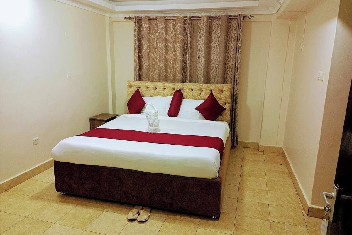 A hotel room with a large bed, a headboard, a nightstand, and a pair of slippers on the floor.