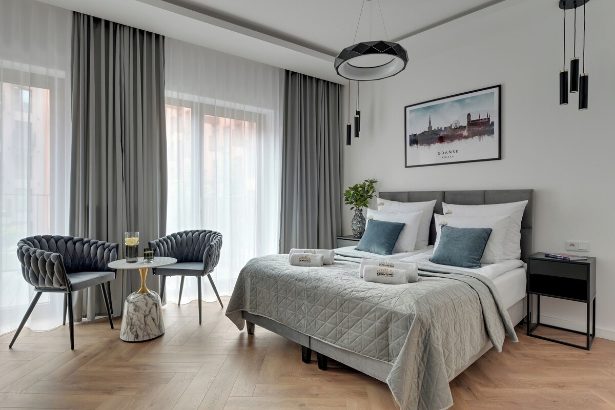 A modern hotel room with a bed, two chairs, a small table, and a cityscape painting.