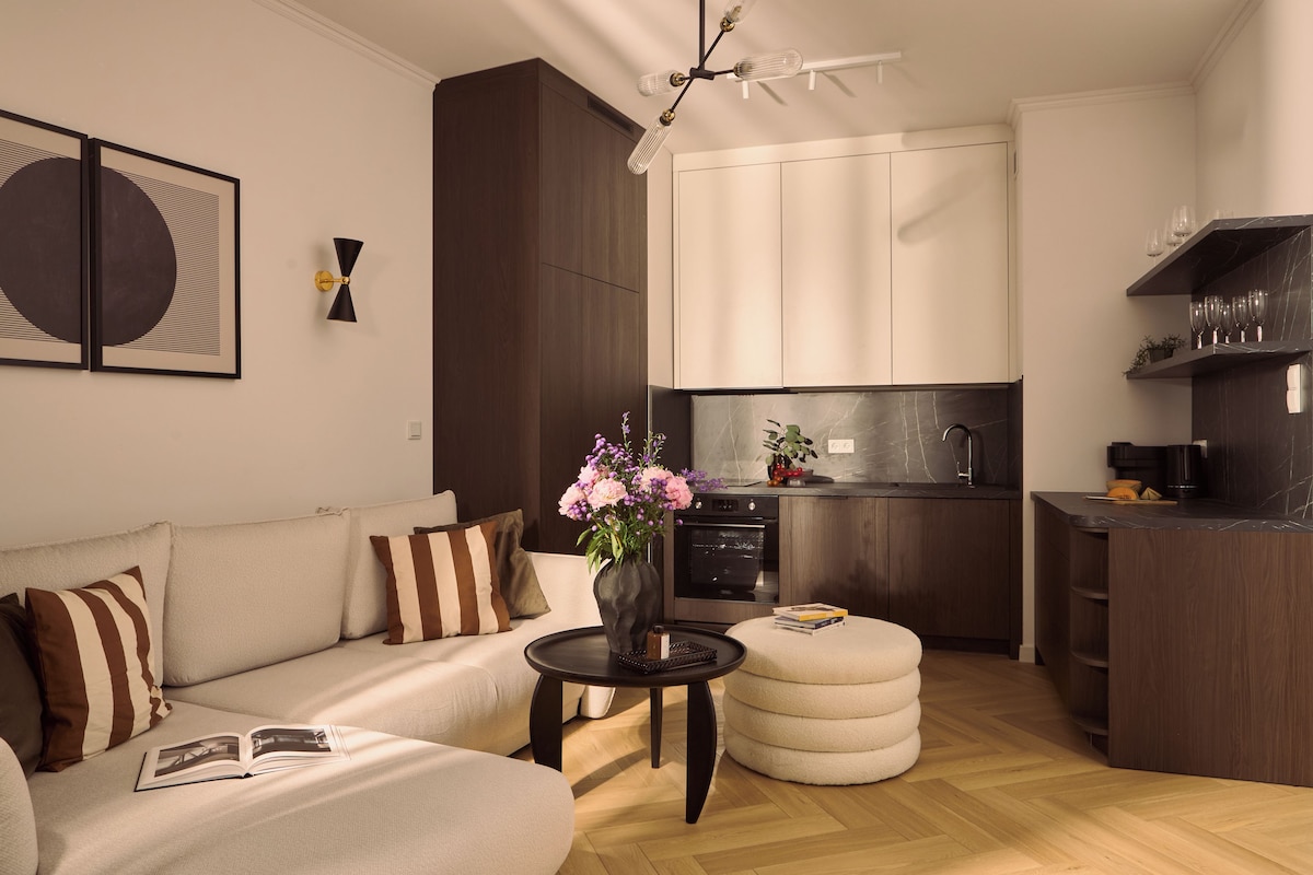A modern living room with a sofa, a coffee table, and a small ottoman. There is a kitchen area with a stove and a sink.