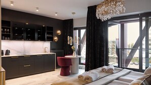 Ein modernes Hotelzimmer mit einem großen Bett, einer kleinen Küchenzeile und Blick auf die Stadt durch große Fenster.