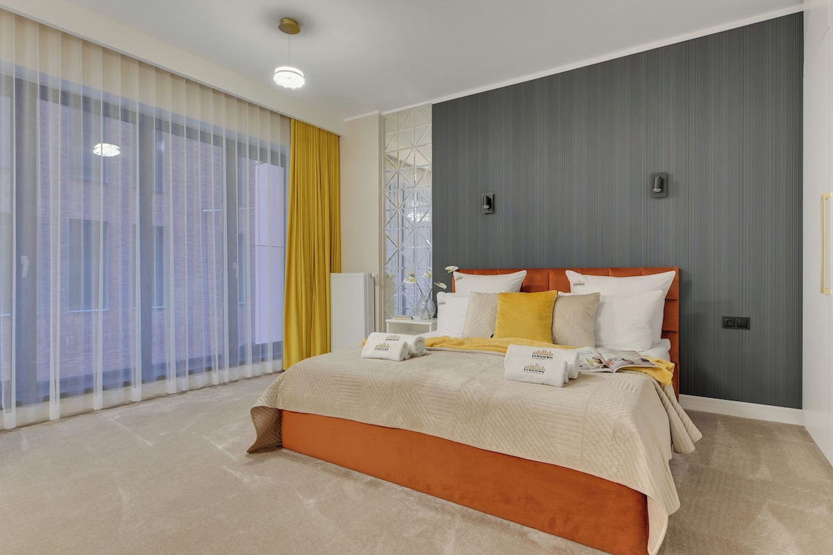 A modern bedroom with a large bed, a bedside table, a window with sheer curtains, and a wall-mounted light fixture.