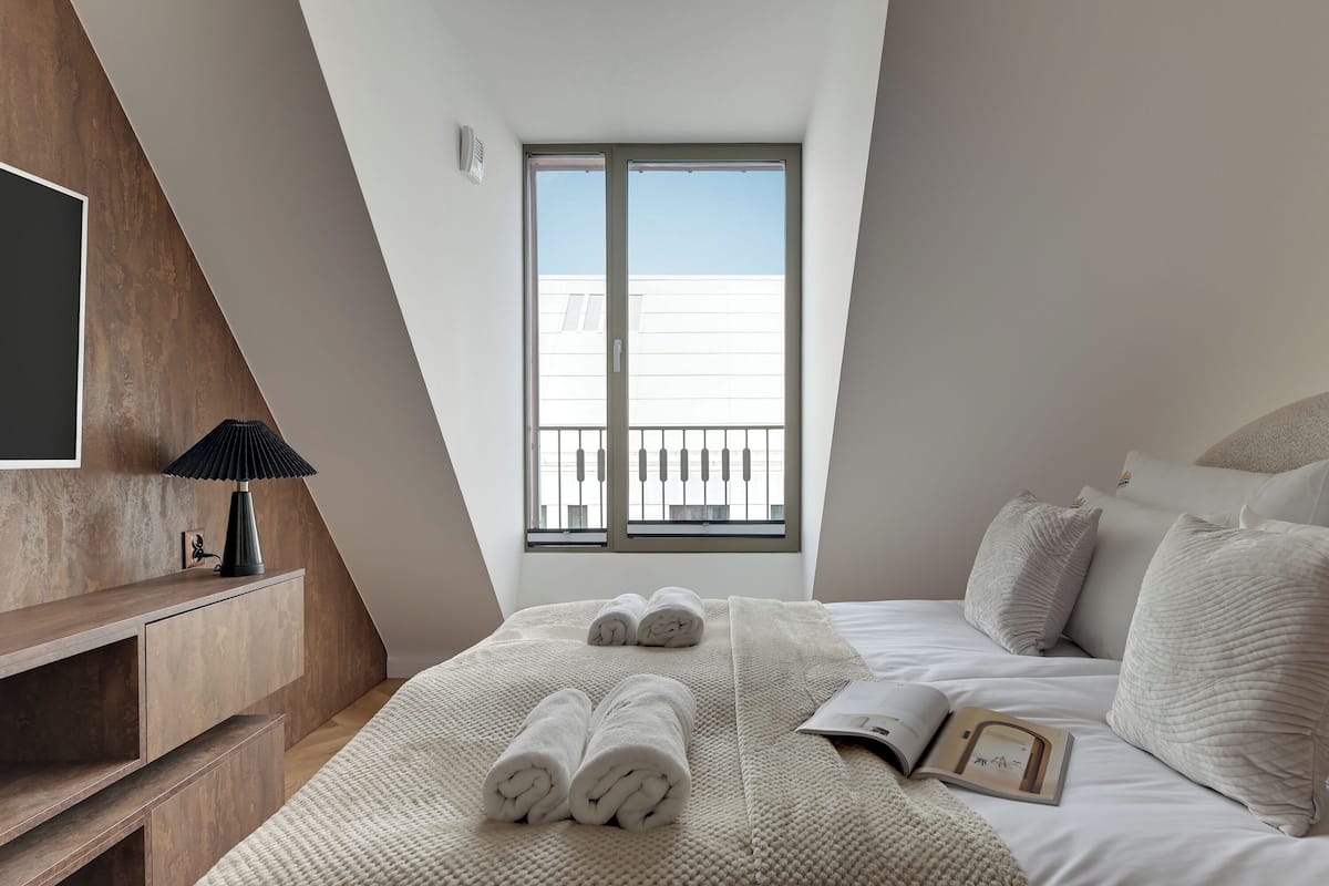 Une chambre avec une grande fenêtre, un lit avec une literie blanche, une table de chevet avec une lampe et une console en bois.