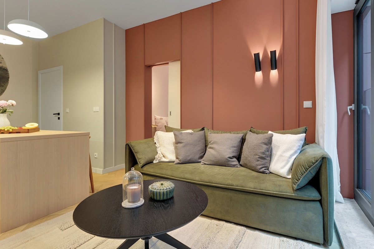 A modern living room with a green sofa, a round coffee table, and a small kitchen area with a fruit tray.