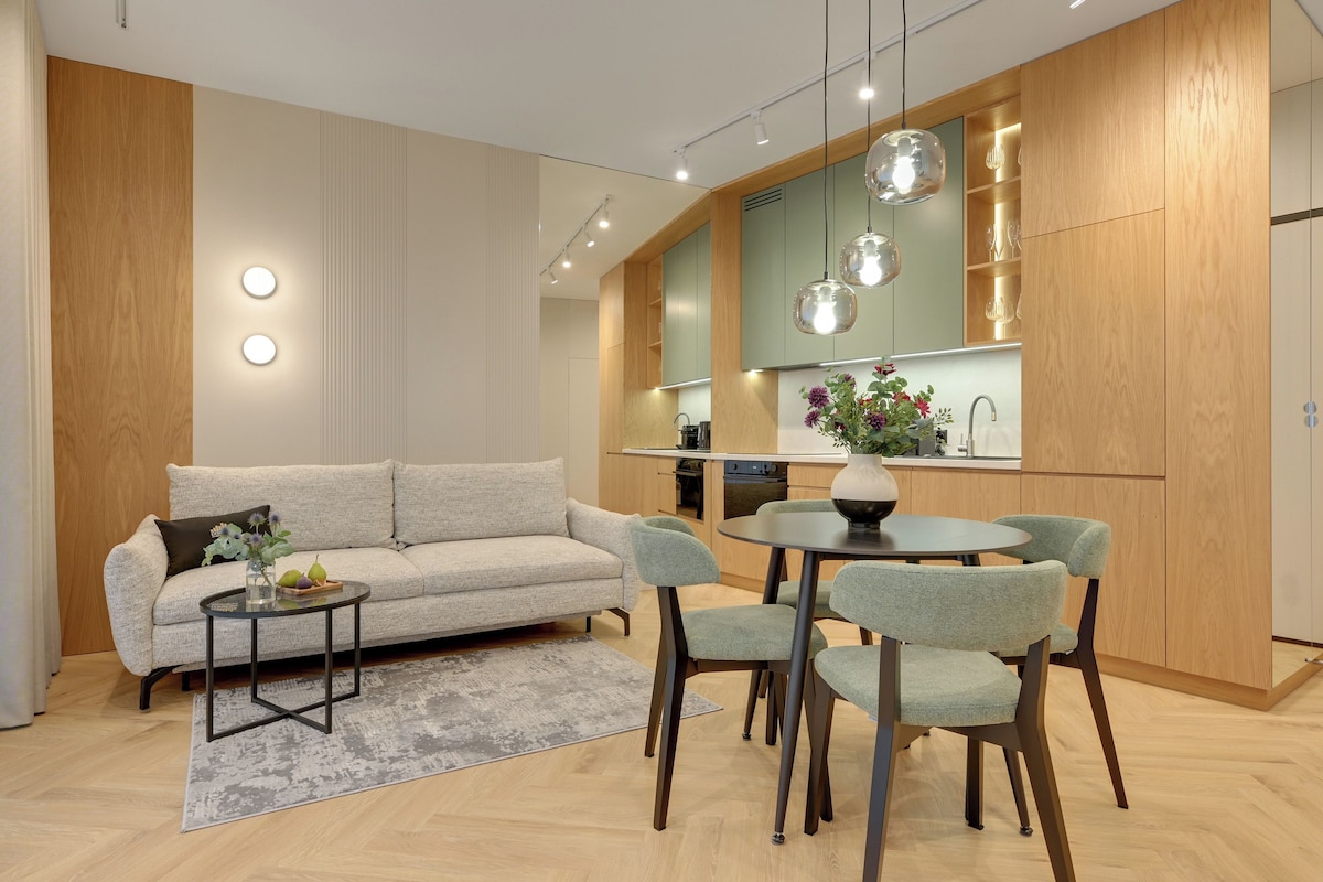 A modern living room with a sofa, coffee table, and chairs. A kitchen with wooden cabinets and a dining area with a table and chairs is visible.