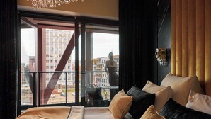 Ein modernes Hotelzimmer mit einem großen Bett, vielen Kissen, Blick auf die Stadt durch große Fenster und einem Balkon mit Geländer.