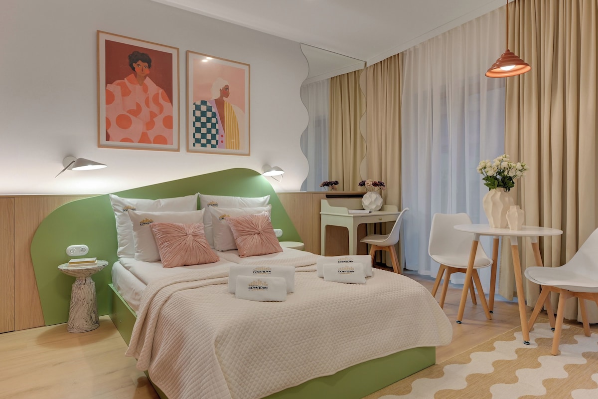 A modern bedroom with a green headboard, a bed with white bedding, a desk, two chairs, and a small table with flowers.
