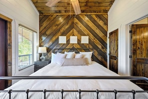 Un dormitorio con techo de madera en espiga, una cama con ropa de cama blanca, una mesita de noche con lámpara y una ventana con persianas.