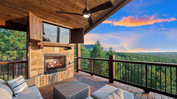 A wooden deck with stone fireplace, outdoor seating, and a scenic forest view.