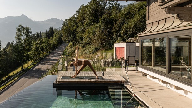 Una persona realiza una postura de yoga sobre una terraza de madera al lado de una piscina cristalina con vistas a la montaña.