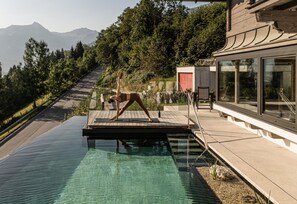 A person performing a yoga pose on a wooden deck by a clear pool with a mountain view.