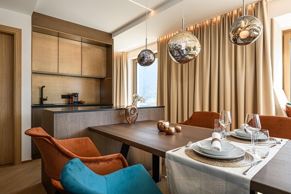 A modern dining area with a wooden table set for two, a kitchen in the background, and large windows with curtains.