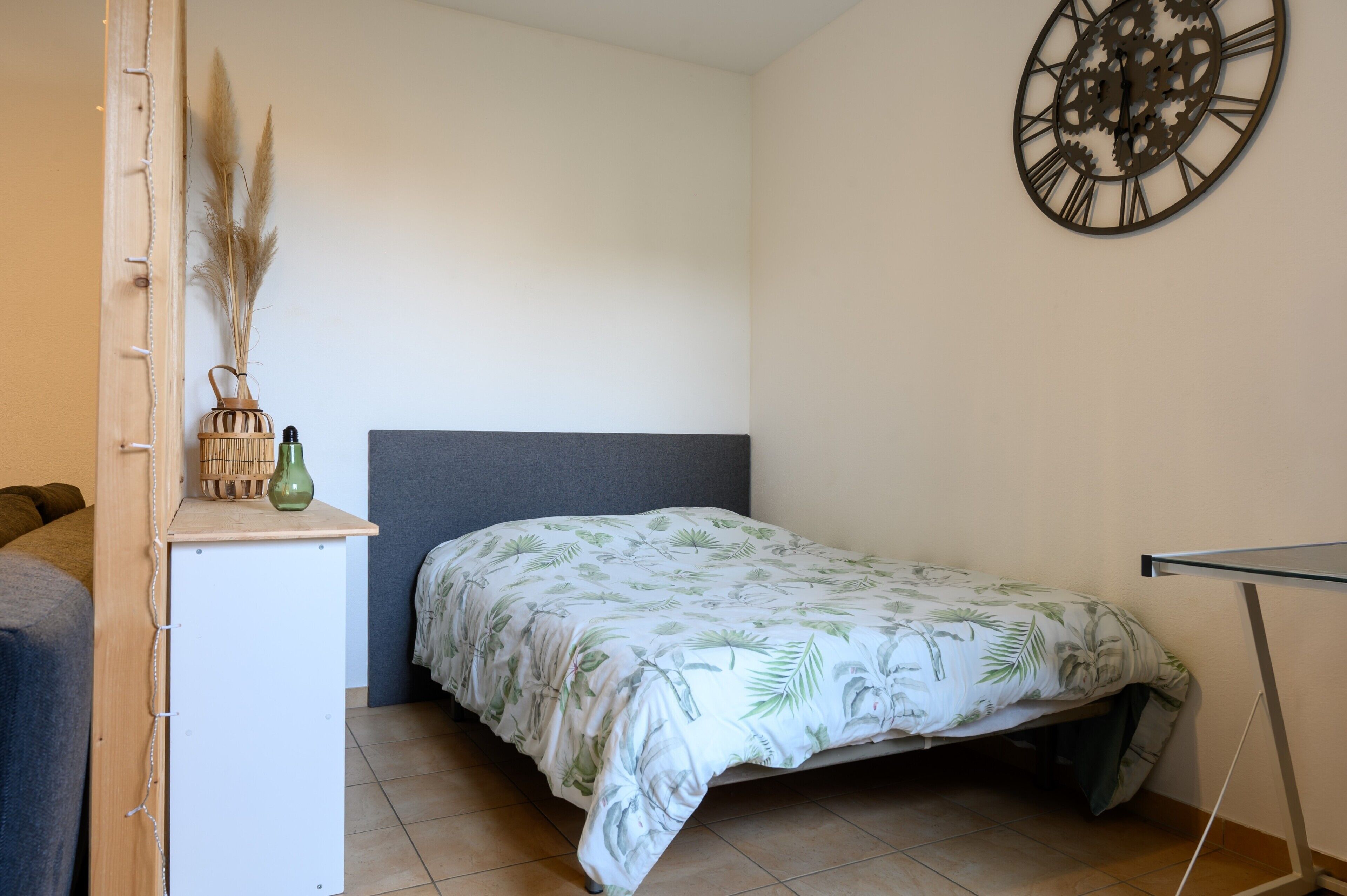 A small room with a bed, a wall clock, and a small table with decorative items.