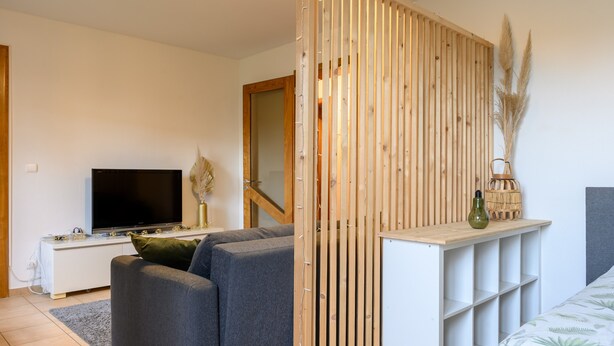 Een moderne woonkamer met een flatscreen televisie, grijze banken, een houten roomdivider en een witte console tafel.