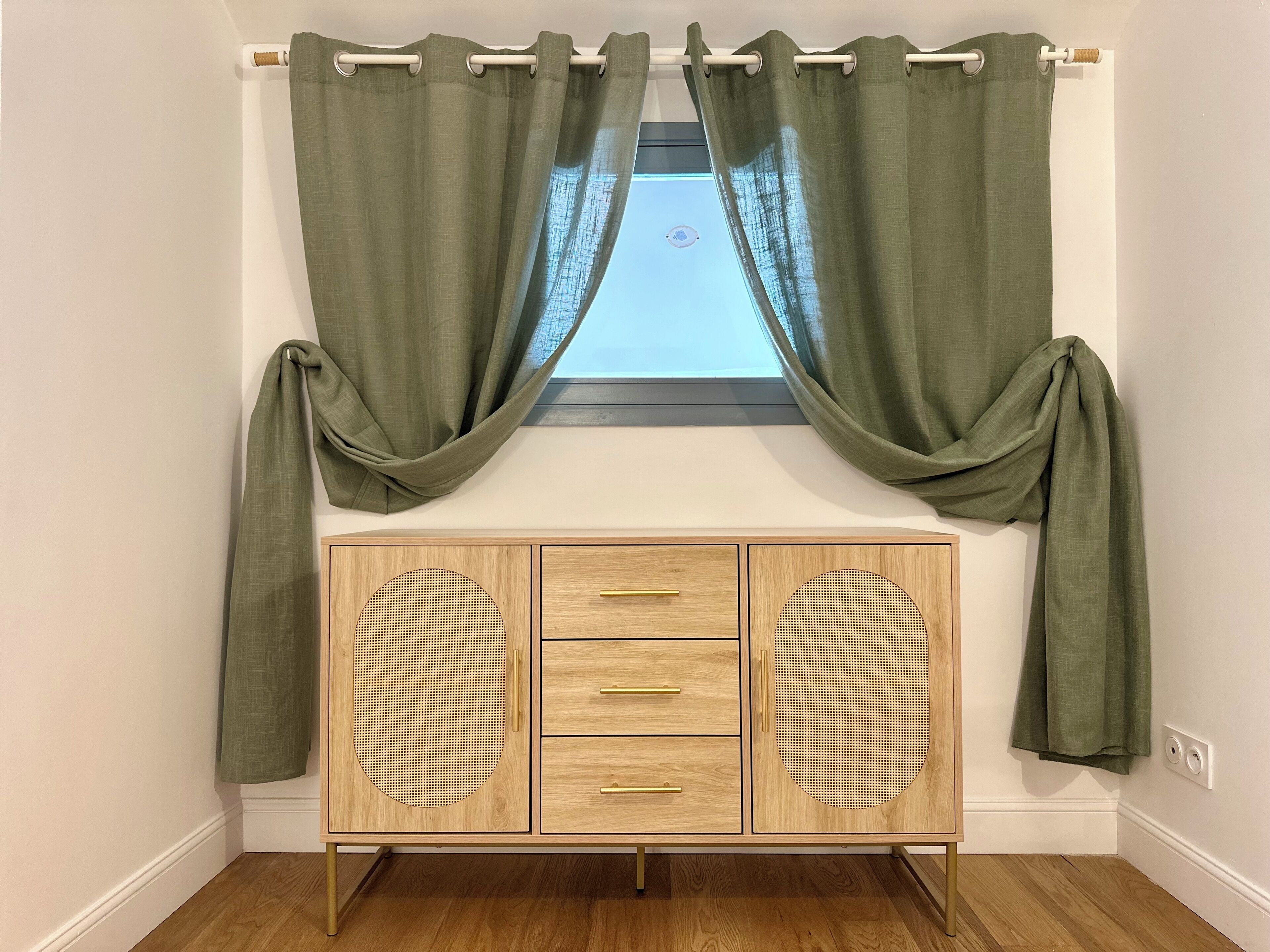 A room with a window, green curtains, a wooden cabinet with drawers, and a wooden floor.